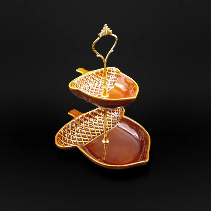 Zweistufige Etagere in Eichelnussform aus braun-orange glasierter Keramik mit goldfarbenem Metallständer und dekorativem Griff, vor schwarzem Hintergrund.