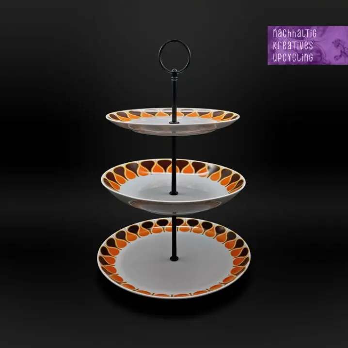 Dreistufige Upcycling-Etagere mit schwarzem Metallstab und Ringgriff; drei weiße Teller mit orange-braunem Retro-Muster am Rand vor schwarzem Hintergrund; oben rechts Schriftzug „nachhaltig kreatives upcycling“.