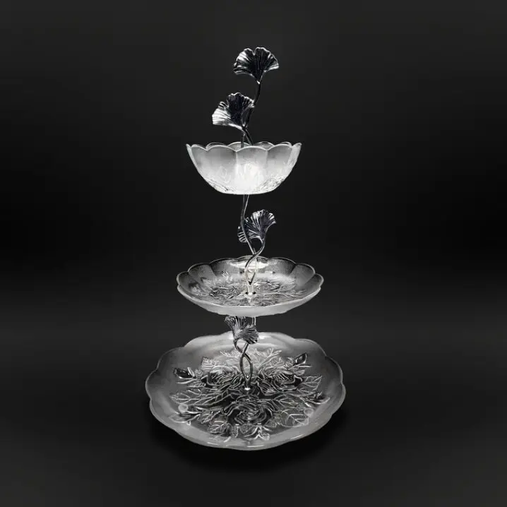 Dreistufige Glas-Etagere mit floral geformten Tellern und dekorativer Metallstange mit Blättern, vor dunklem Hintergrund.