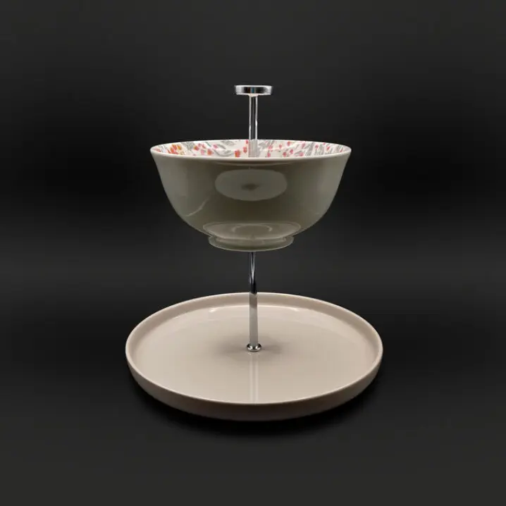 Zweistufige Etagere mit silberfarbenem Metallständer: oben eine Schale mit floralem Muster (rosa/orange Blüten), unten ein beigefarbener Teller, vor schwarzem Hintergrund.